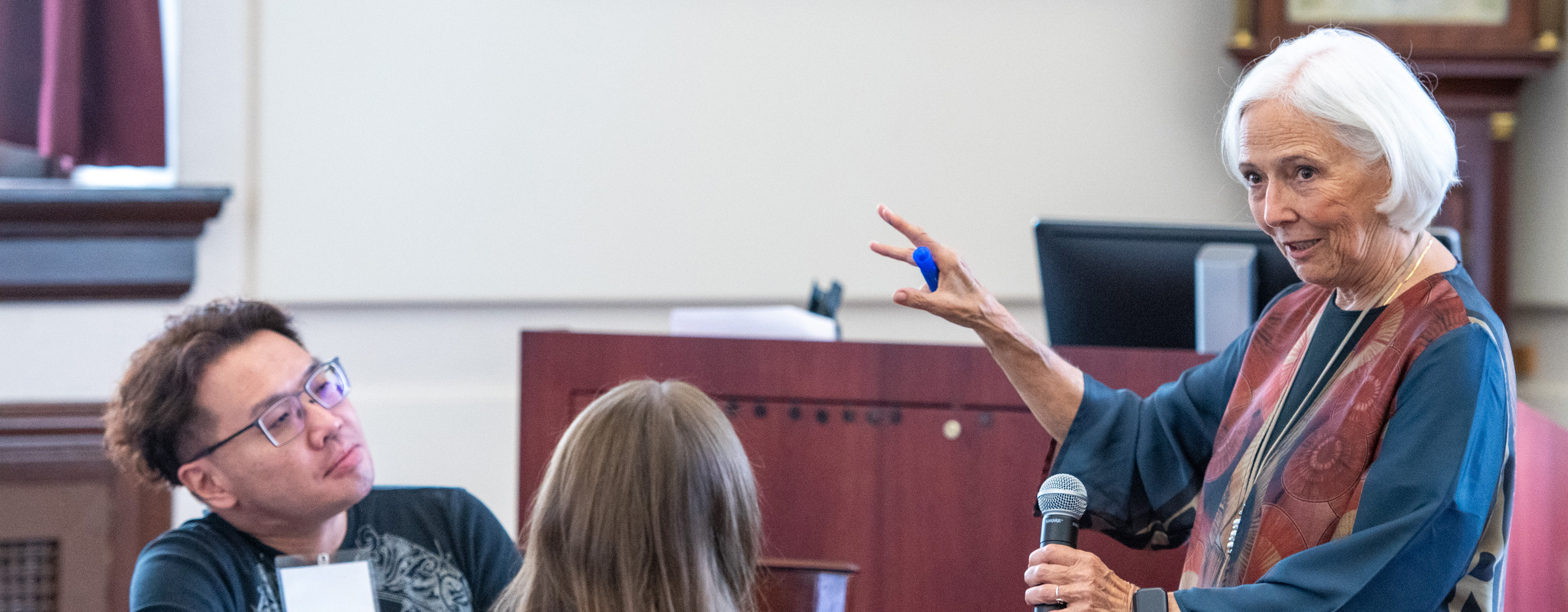 Professor with short grey hair and colorful dress speaks into microphone.