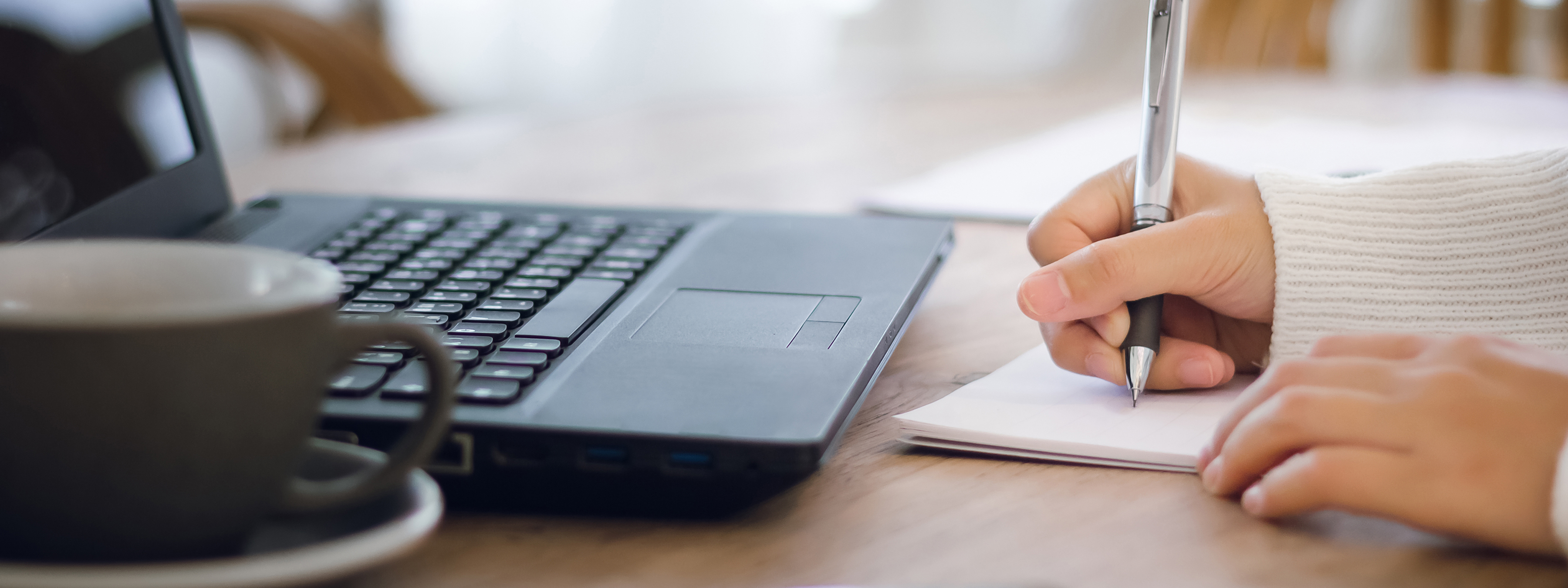 Hand holding pen writing in journal beside open laptop and coffee mug.