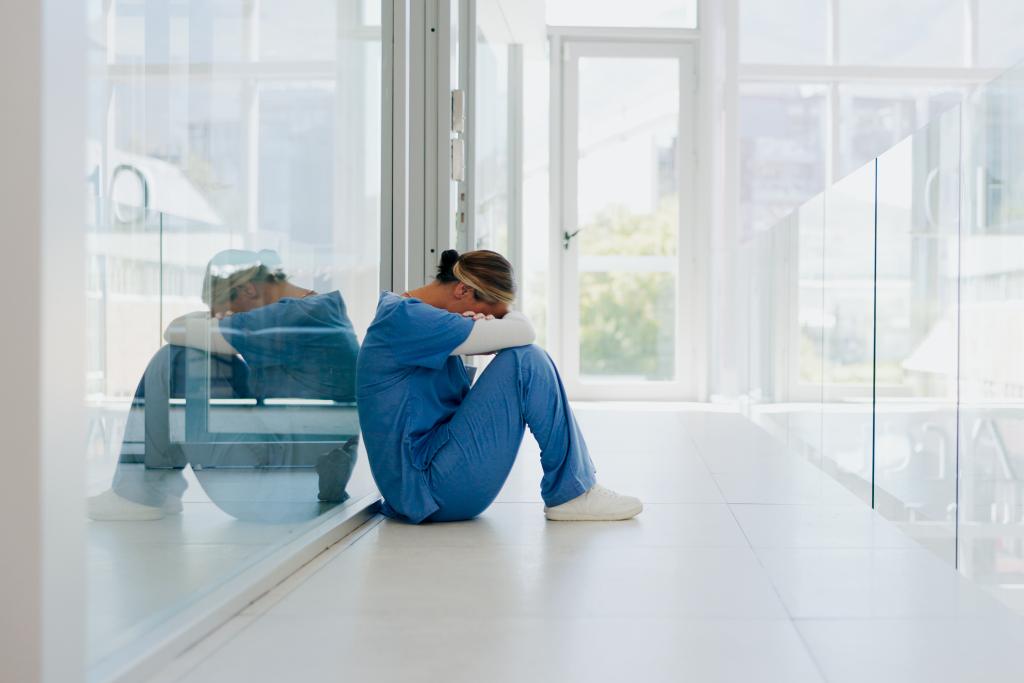 Overworked healthcare worker wearing blue scrubs sits on hospital floor, resting her head on her knees.