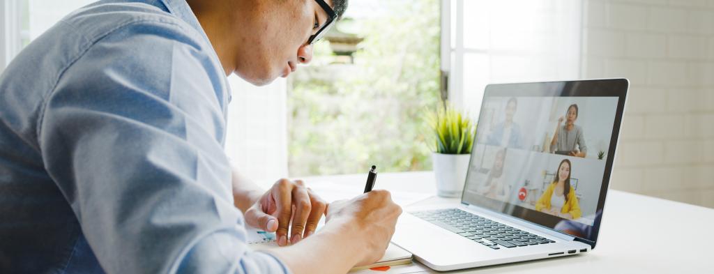 Young man writing in a notebook while in a virtual meeting.