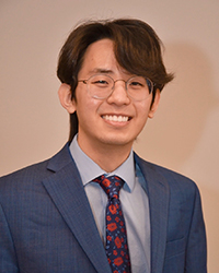 William Choi smiling, wearing round glasses, a blue suit, and a floral pattern tie
