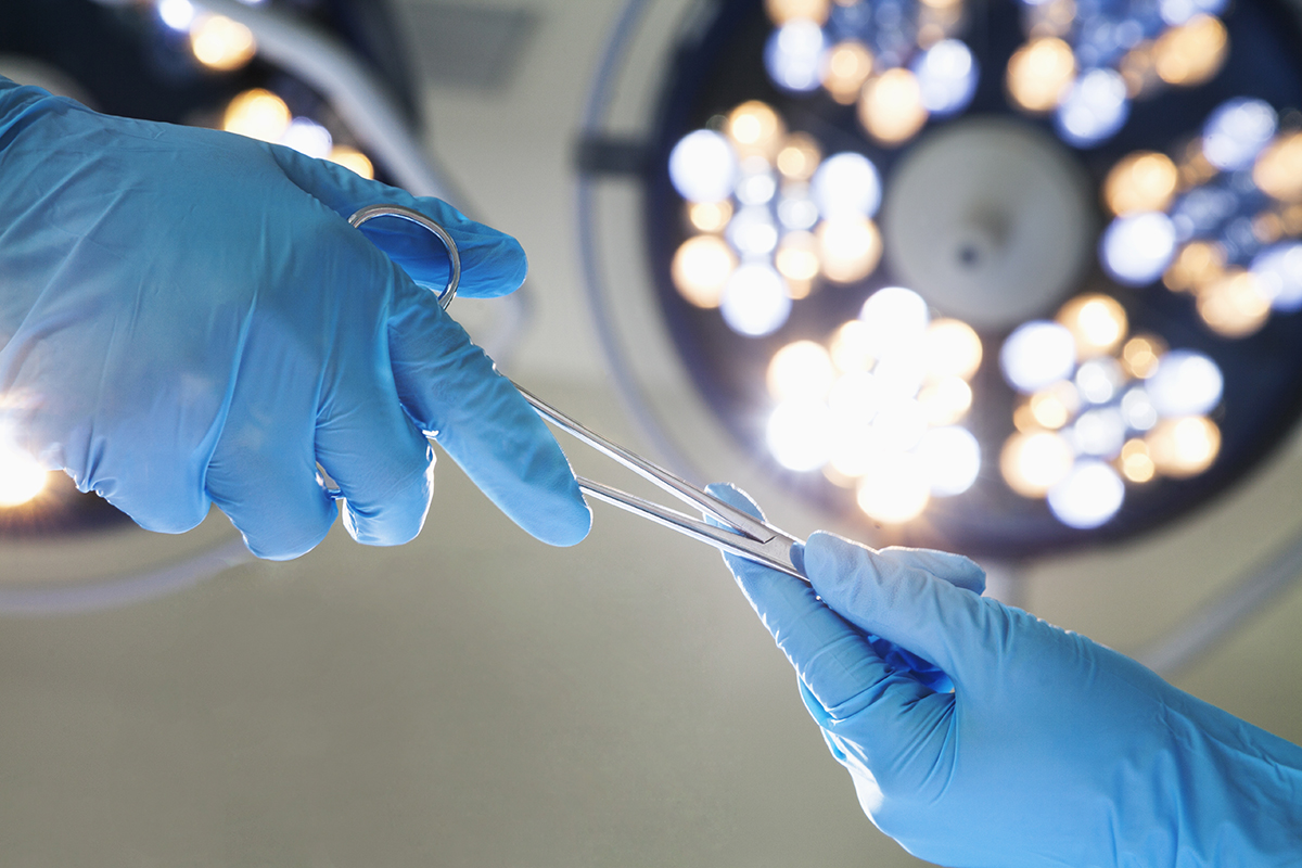 Two gloved hands handing off a surgical instrument under a bright operating light