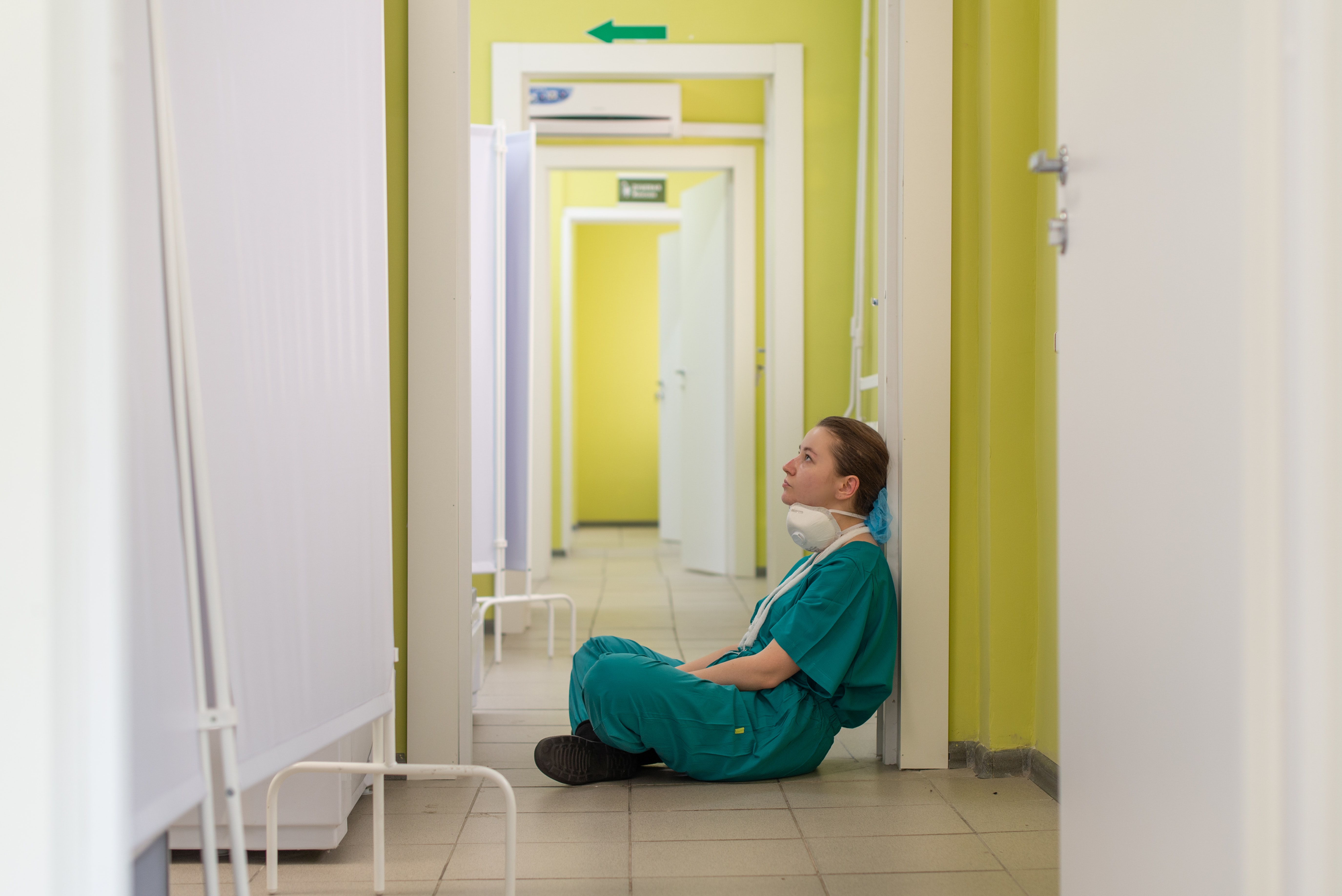 A tired nurse wearing teal scrubs sits on the floor of a hospital